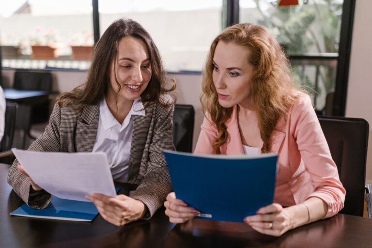 Dwie kobiety siedzące za biurkiem i analizujące trzymane w rękach dokumenty