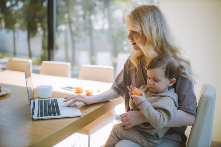 Kobieta z dzieckiem na kolanach pracująca przy laptopie w domu